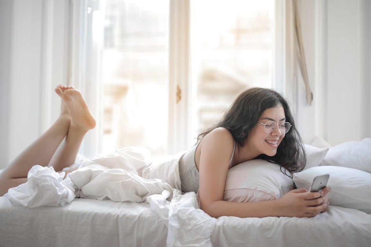 Smiling woman on her phone in bed, enjoying a virtual date with sex toys for long distance couple.