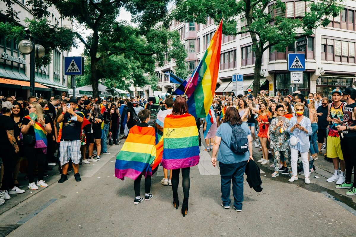 Colorful pride parade with participants wrapped in rainbow flags, walking through a crowded city street filled with spectators and celebratory energy.