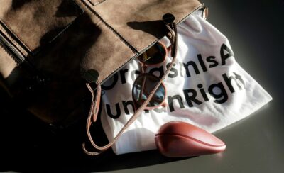 Flat lay of a stylish brown leather bag, a pair of sunglasses, a white t-shirt with bold text partially visible, and a metallic pink accessory, all arranged on a dark surface with natural lighting.