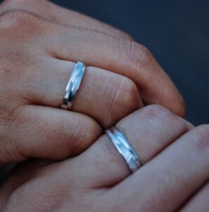 Close-up of two hands wearing matching silver wedding bands, symbolizing love and commitment, with a blurred dark background.