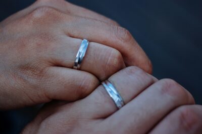 Close-up of two hands wearing matching silver wedding bands, symbolizing love and commitment, with a blurred dark background.