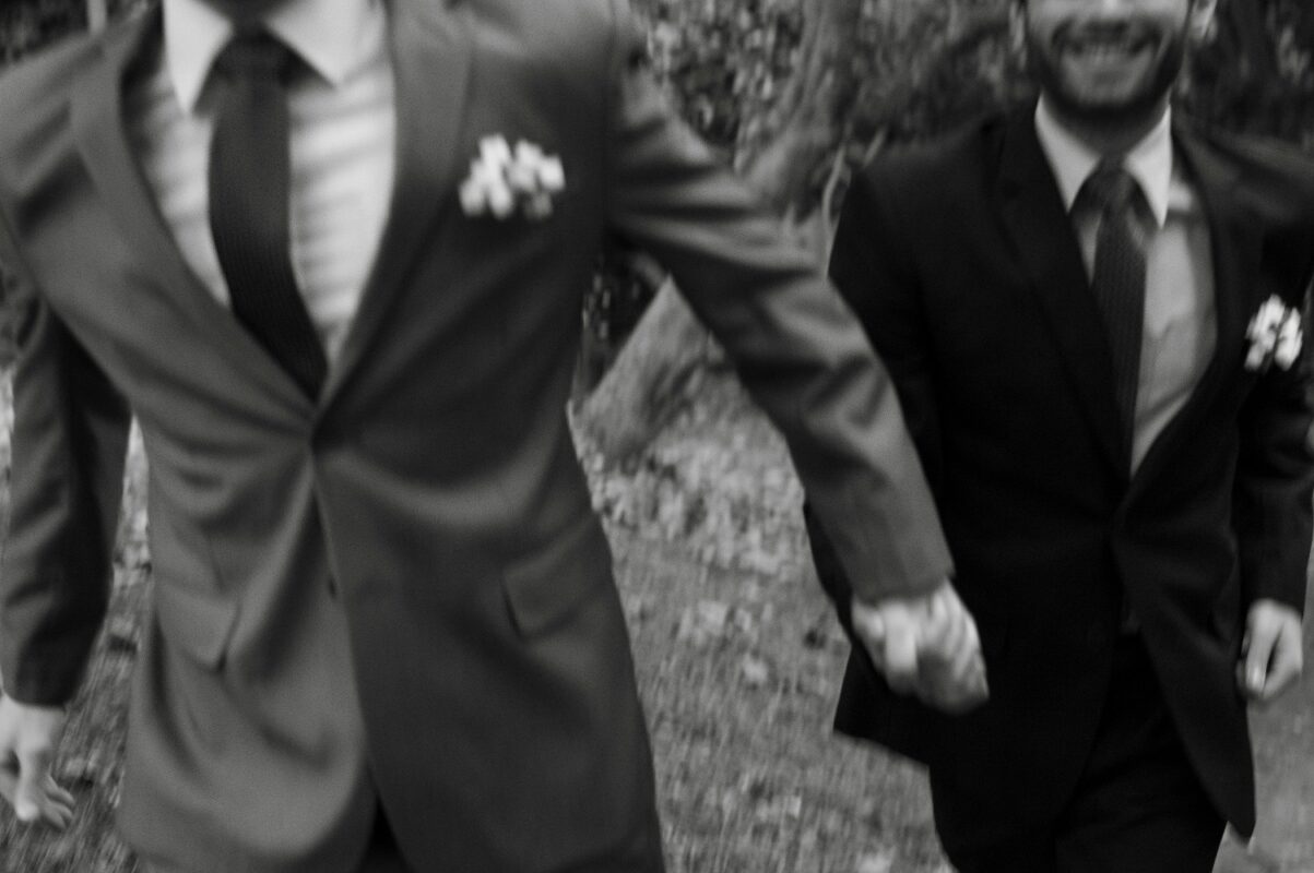 Black-and-white image of two grooms in suits holding hands and smiling as they walk together, symbolizing love and unity.