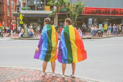 Two individuals wrapped in rainbow flags standing on a street during a pride event.