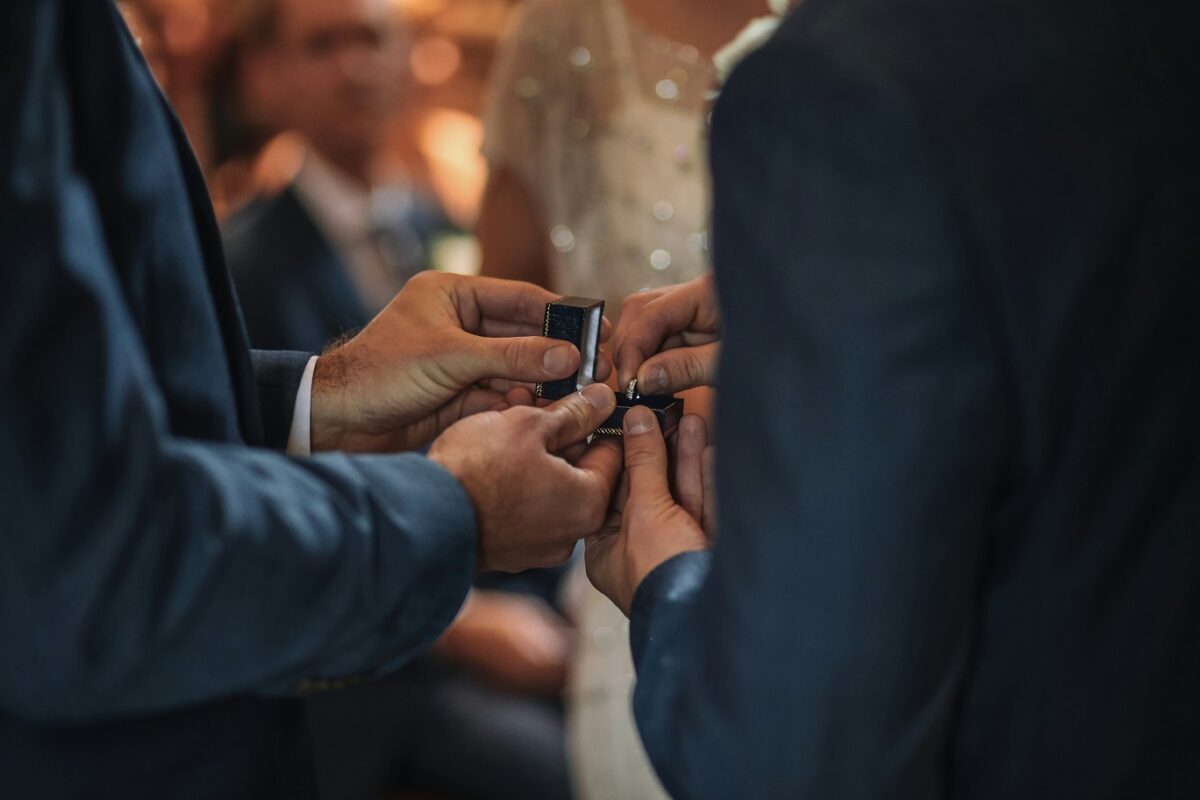 Close-up of two individuals exchanging rings during a wedding ceremony, with hands holding ring boxes and a blurred audience in the background.