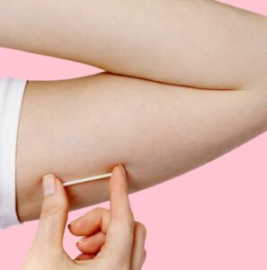 Close-up of a person inserting a Nexplanon birth control implant into the upper arm against a pink background