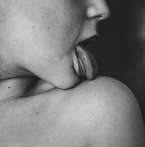 A black and white photo capturing a person’s tongue sensually touching a shoulder, expressing intimacy and erotic tension.