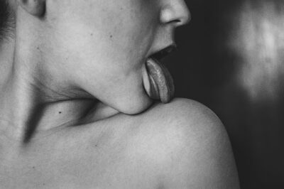 A black and white photo capturing a person’s tongue sensually touching a shoulder, expressing intimacy and erotic tension.