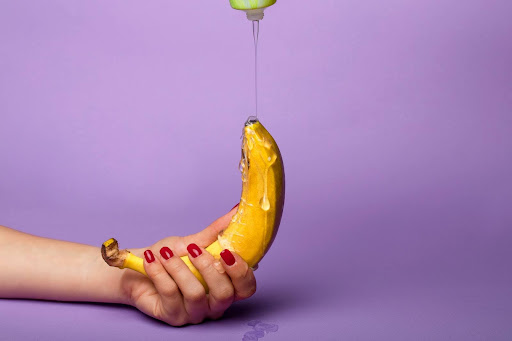 A peeled banana held by a hand with red nails as lubricant drips from above, set against a purple background for a sensual metaphor.