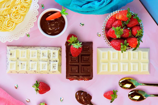 Assorted white and dark chocolate bars with strawberries and chocolate dip arranged on a pink surface for a playful dessert flatlay.