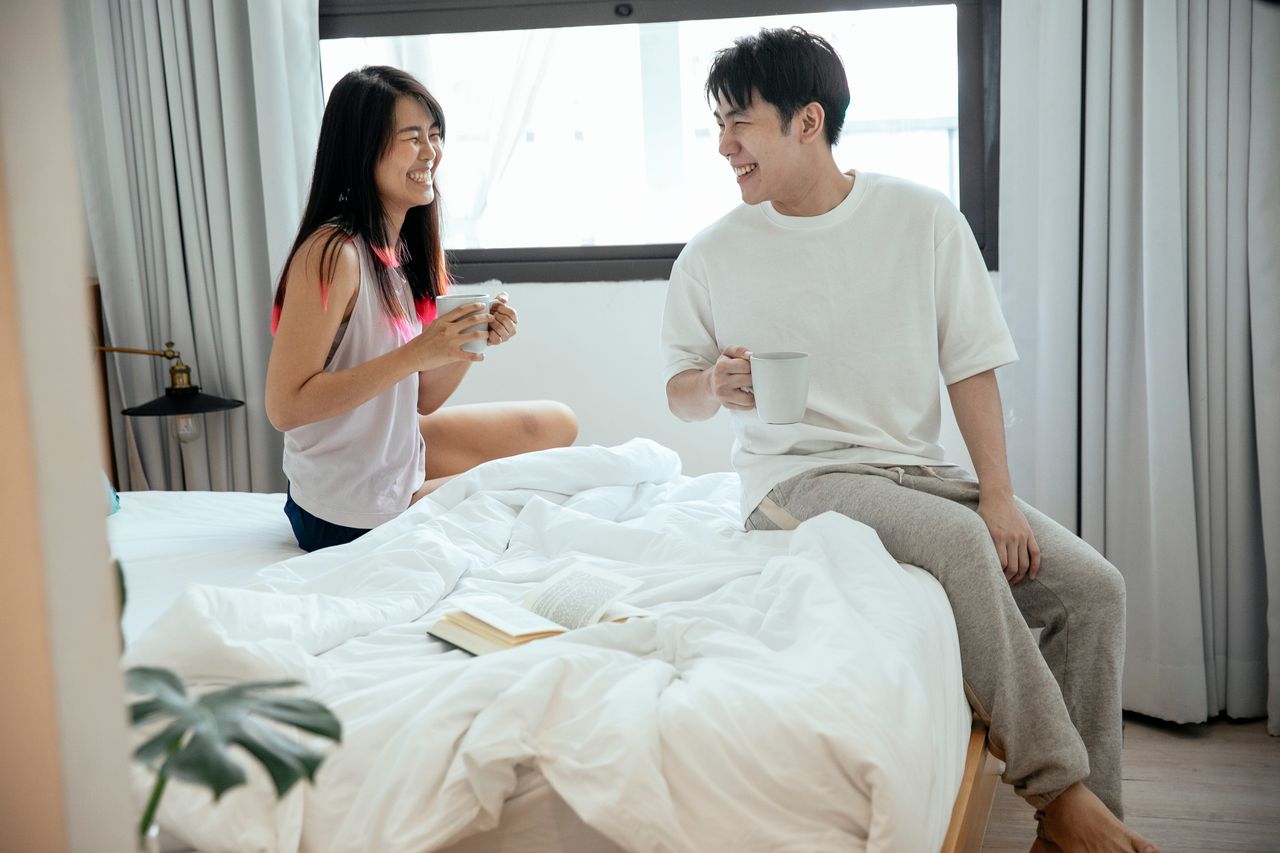 Couple laughing together in bed with morning coffee, reflecting healthy intimacy and open communication as part of intimate wellness. 
